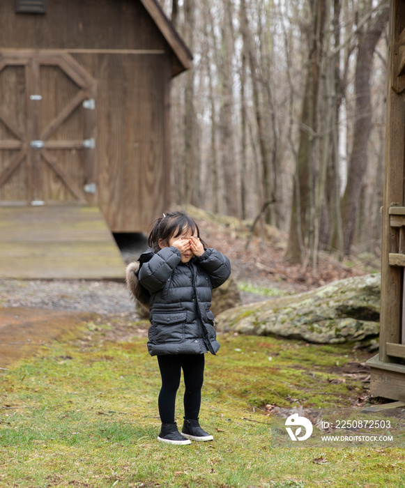 Girl with hands covering eyes standing on grassy field against cottage in forest