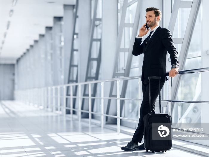 Happy wealthy businessman standing in airport, talking on mobile phone