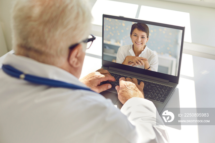 Mature male doctor sitting at desk in office giving video consultation to happy woman