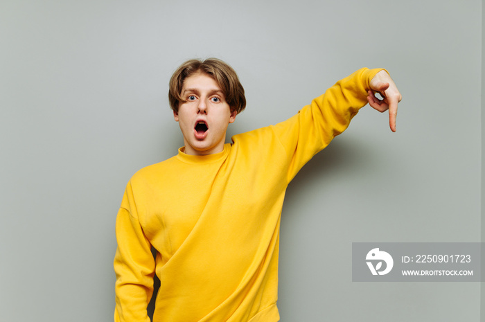 Surprised young man stands on a gray background with a shocked face and shows his finger down on a b