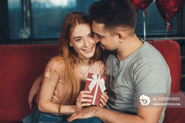 Caucasian couple, girl holding in hands red gift box for anniversary congrats sitting on sofa. Happy