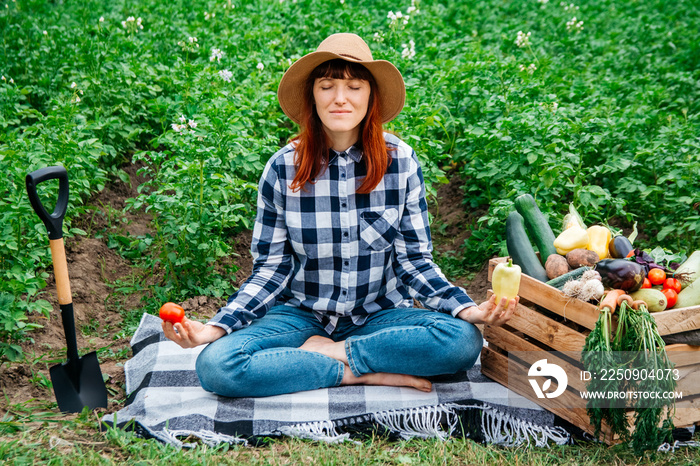 Beautiful female farmer meditating while sitting on a blanket near fresh organic vegetables in a woo