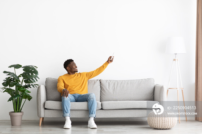 Joyful african american guy sitting on couch, turning on AC