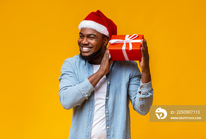 Happy african guy shaking Christmas gift box, listening what rattles