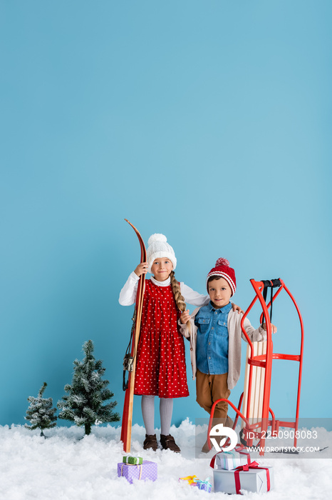child in winter outfit holding skis and hugging boy standing near sleight isolated on blue