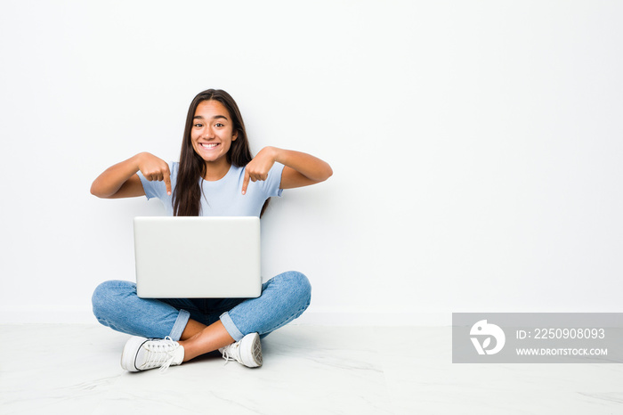 Young mixed race indian woman sitting working on laptop points down with fingers, positive feeling.