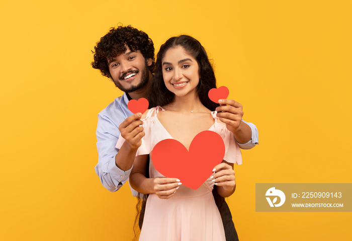 Love is in the air. Romantic indian couple with red paper hearts in hands posing over yellow studio 