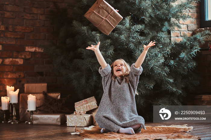 Joyful little girl is tossing her gift while sitting near christmas tree.