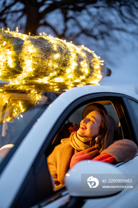 Happy woman driving car with a illuminated Christmas tree on a rooftop. Concept of New Year preparat