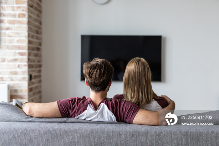 一对幸福的热恋情侣坐在家里的沙发上看电视的背影。