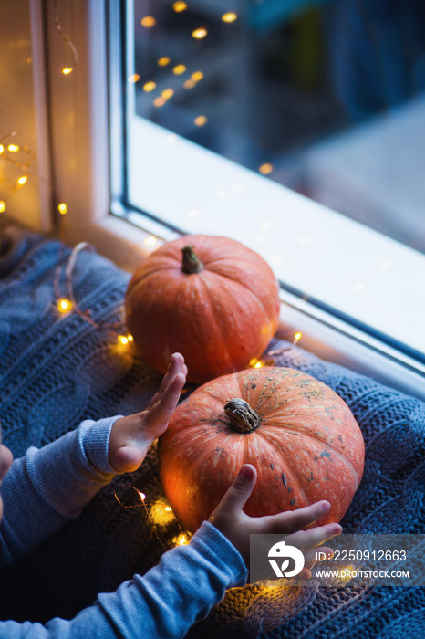 Toddler boy holding orange pumpkin on gray knitted plaid near window in evening surrounded with warm