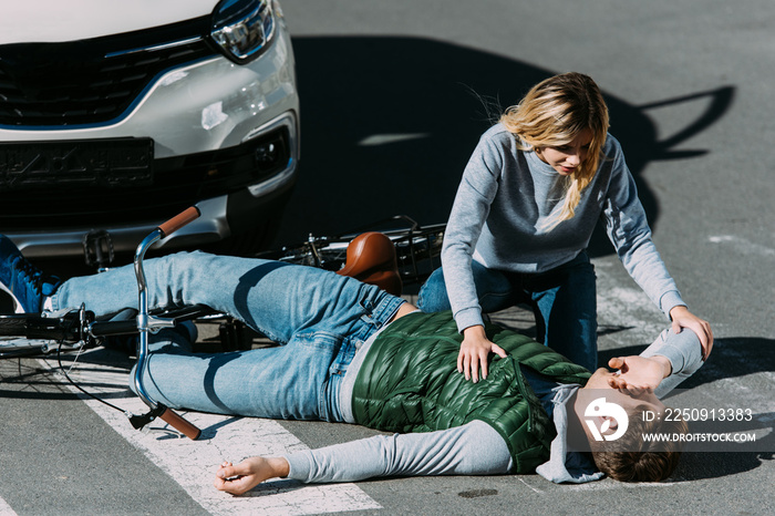 woman looking at injured young cyclist lying with bicycle on road at car accident