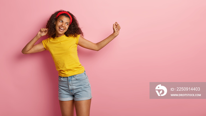 Horizontal view of pleased girl with Afro haircut spreads hands, dances against pink background, foc