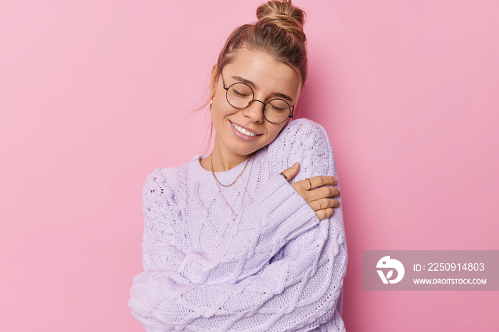 Beautiful pleased young woman with fair hair gathered in bun embraces own body wears knitted sweater