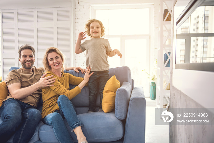 Smiling parents are embracing on cozy couch and looking at camera with joy. Joyful son is standing o