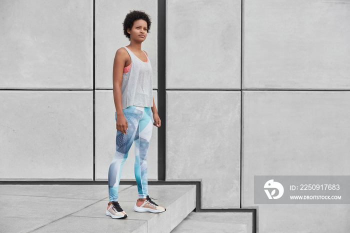 Horizontal full length shot of self determined thoughtful female fitness trainer stands at stairs, w