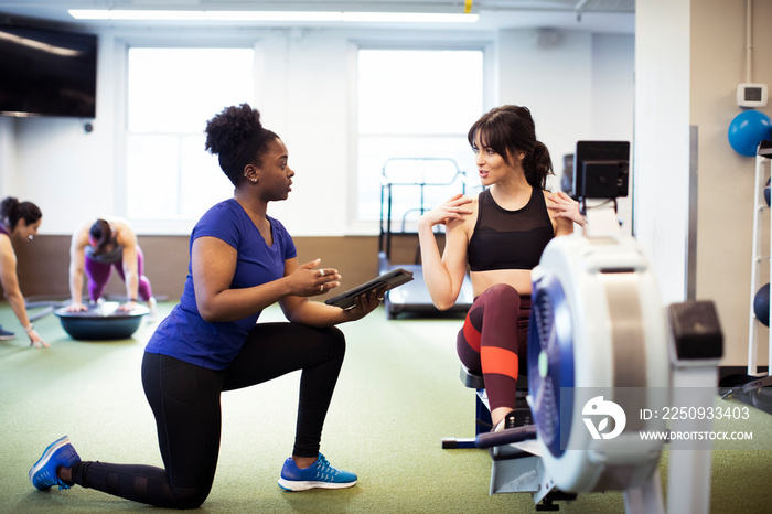 Instructor holding tablet computer discussing with female athlete exercising on rowing machine at gy