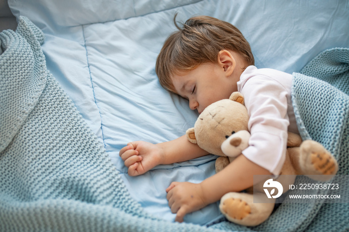 Healthy child, sweetest blonde toddler boy sleeping in bed holding her teddy bear. Adorable toddler 