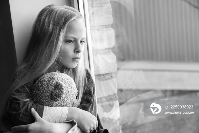 Homeless little girl with teddy bear sitting on window sill