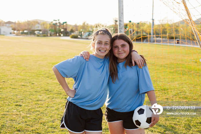 Portrait of smiling girls with ball standing on soccer field against clear sky