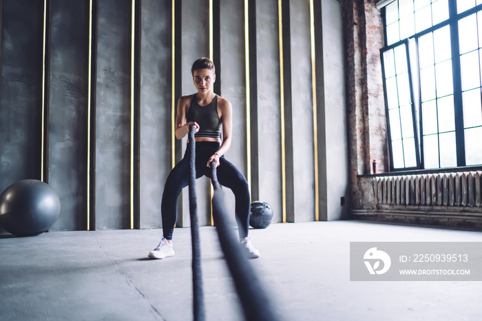 Confident caucasian female in active wear using rope for slimming and working on arms muscles in gym