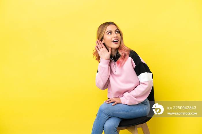 Young woman sitting on a chair over isolated yellow background listening to something by putting han