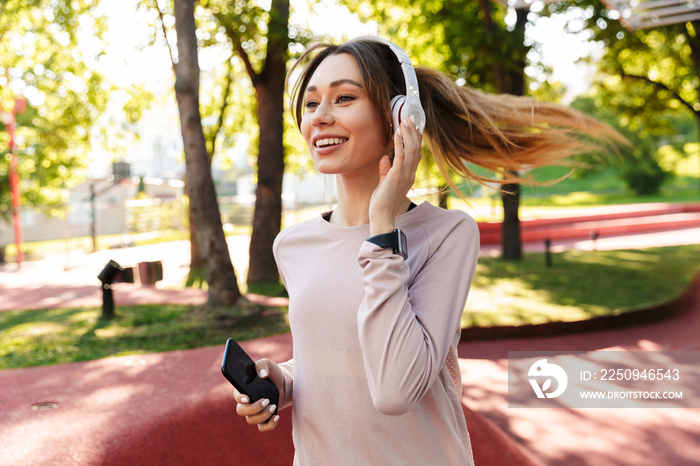 Beautiful cheerful young fitness sports woman posing outdoors in park listening music with earphones