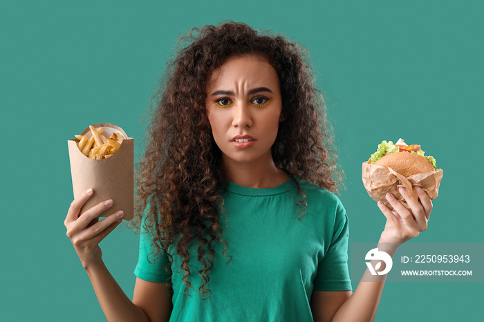 Sad young African-American woman with french fries and burger on color background