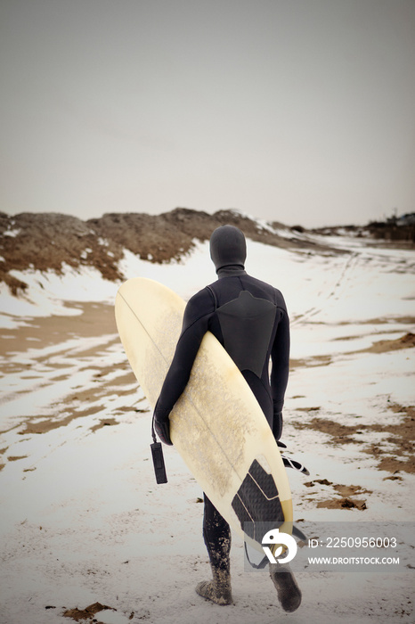 Rear view of man wearing wetsuit walking across along snowy beach carrying surfboard