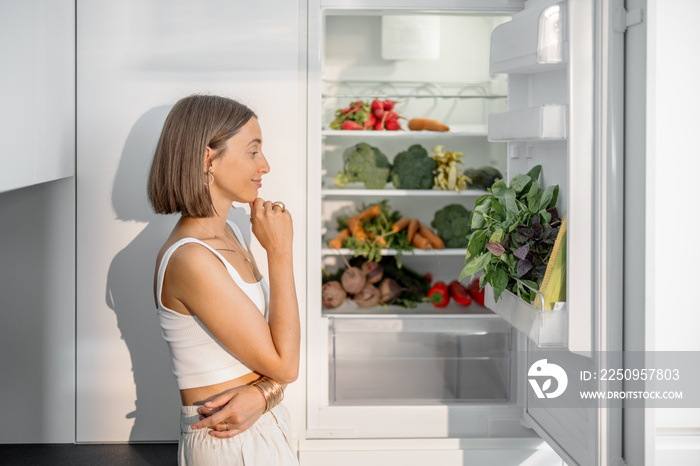 年轻女子站在现代厨房里装满新鲜蔬菜的冰箱旁。健康的素食主义者