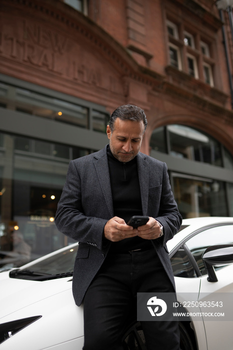 Mature man leaning against luxury car and using phone