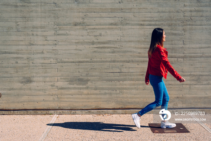 Millenial woman walking calmly in blue and red jeans next to a cement wall, gray background copy spa