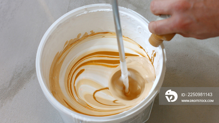 Hand pouring brown liquid into white paint. Person mixing white paint in bucket. Home renovation con