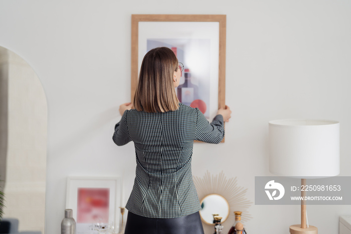 家居装修。一位女士在光线充足的室内白墙上挂着一幅画的后视图