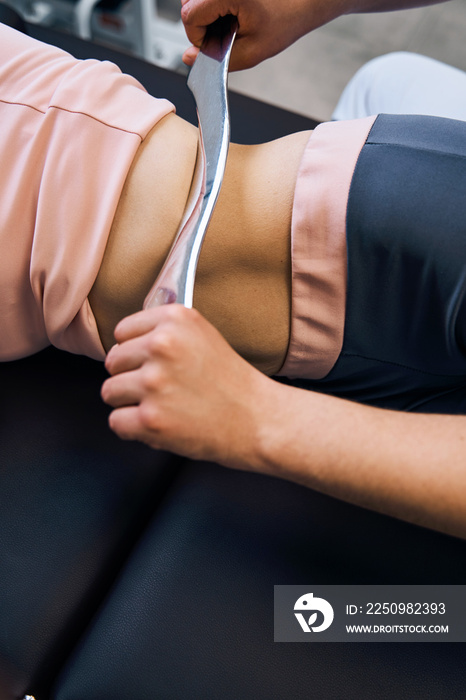 Close up injured young woman lies on therapy bed while physiotherapist massages abdomen with IASTM