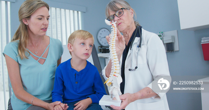 Senior woman pediatrician with spine model talking to little boy patient and his mother. Child looki