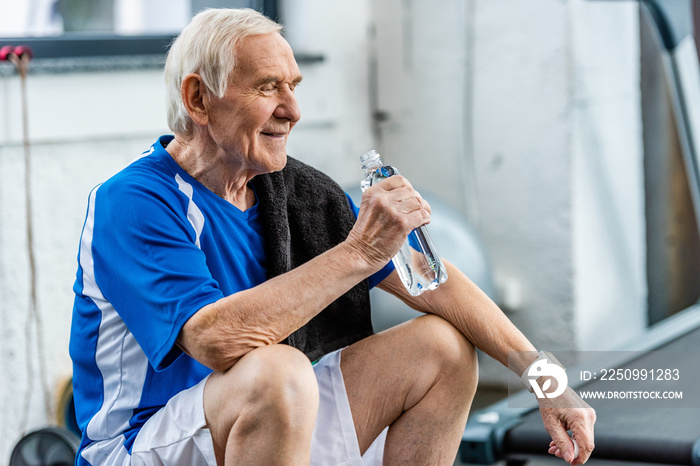 smiling senior sportsman with towel and bottle of water resting at gym