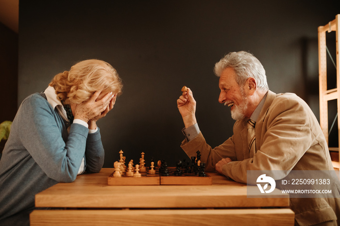 Close photo of elderly couple playing chess at home. Woman is not happy because man is winning.