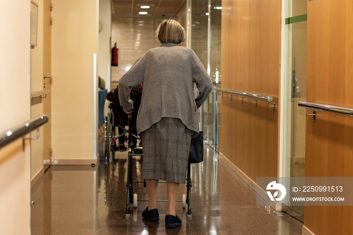 Rear view of old woman walking with roller walker in corridor.