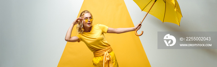 panoramic shot of attractive blonde girl touching sunglasses and holding umbrella on white and yello