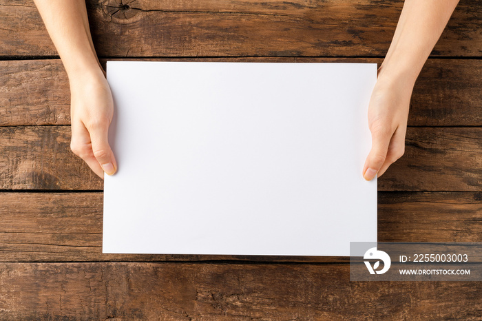 Young woman’s hands showing blank paper sheet on rustic wooden desktop. Top view