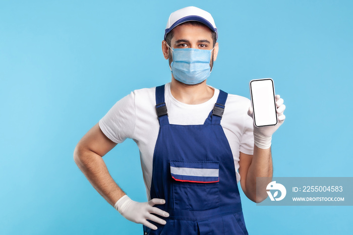 Worker in overalls, mask and gloves holding mobile phone with blank display, device mock up for onli