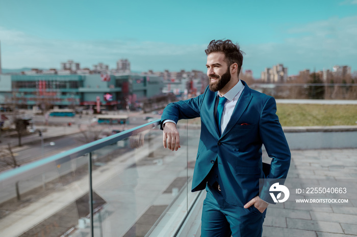 Young prosperous smiling bearded businessman in formal wear standing at rooftop and looking at citys