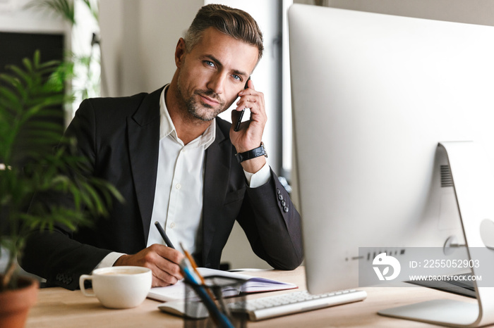 Image of adult businessman talking on cell phone while working on computer in office