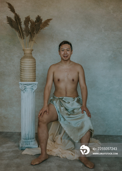 Chinese man with scar on stomach sitting with hands on thighs next to roman column