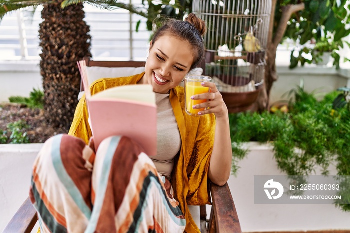 Young hispanic girl reading book and drinking orange juice at the terrace.