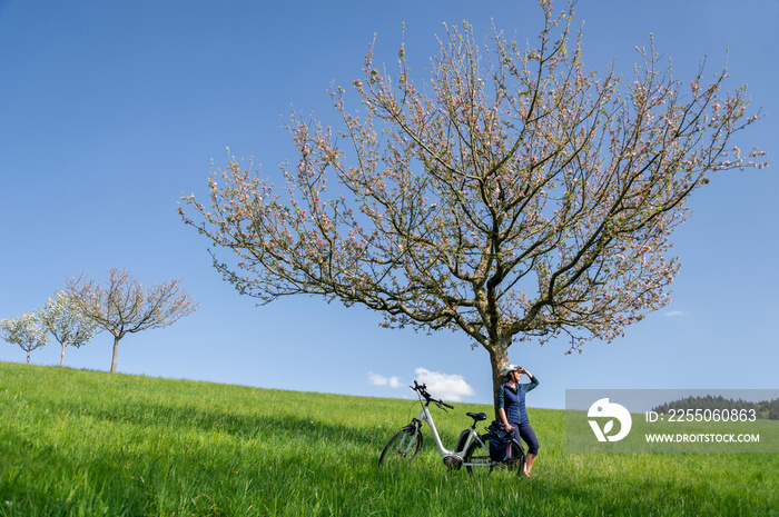 Frau mit Fahrrad an blühenden Apfelbaum gelehnt, Frühlingsstimmung im Odenwald, Sonne, blauer Himmel