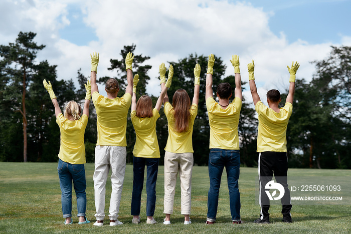 Rear view of happy volunteers wearing yellow uniform and rubber gloves raising hands up, celebrating