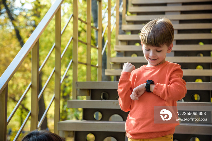 一个穿着时髦衬衫的可爱男孩呆在学校附近，看着他的智能手表触摸屏幕。A