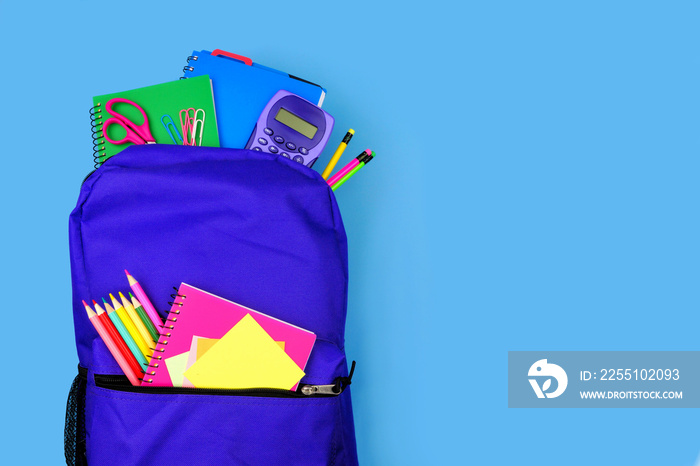 Purple backpack full of school supplies against a blue background. Close up, top view with copy space.